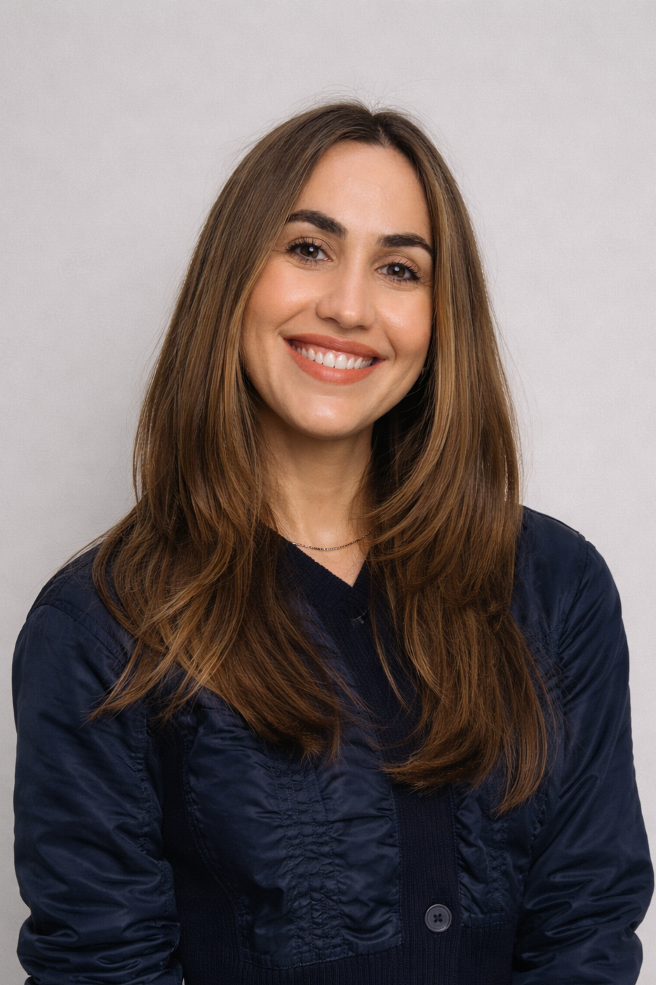 A woman with long brown hair smiling, wearing a black jacket, standing against a light gray background.