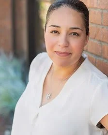 A woman with dark hair pulled back, wearing a white top, smiling slightly, standing outdoors near a brick wall.