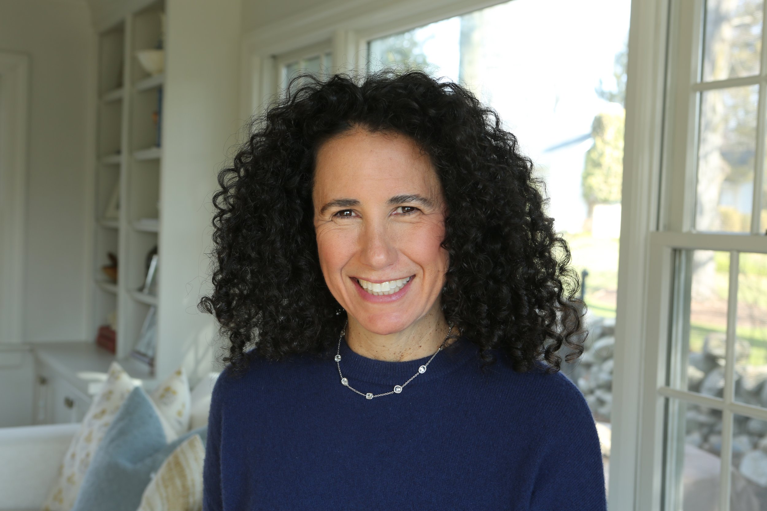 A woman with curly black hair smiling indoors near a large window with trees outside.