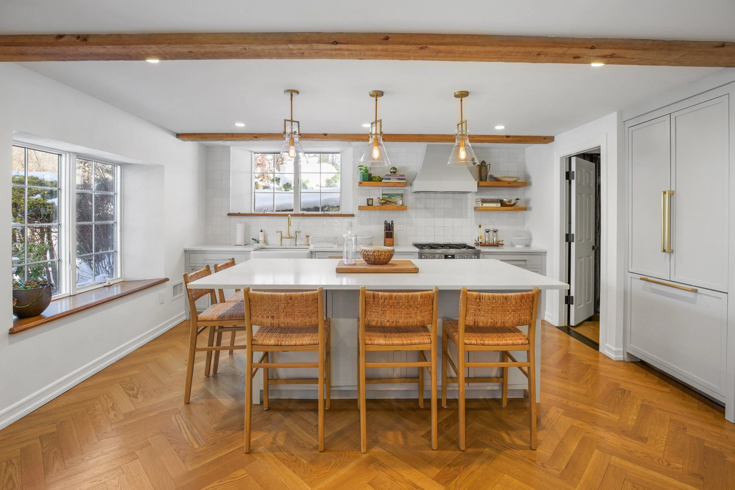Bright kitchen with large central island, wooden chairs, open shelving, and a window with snow outside.