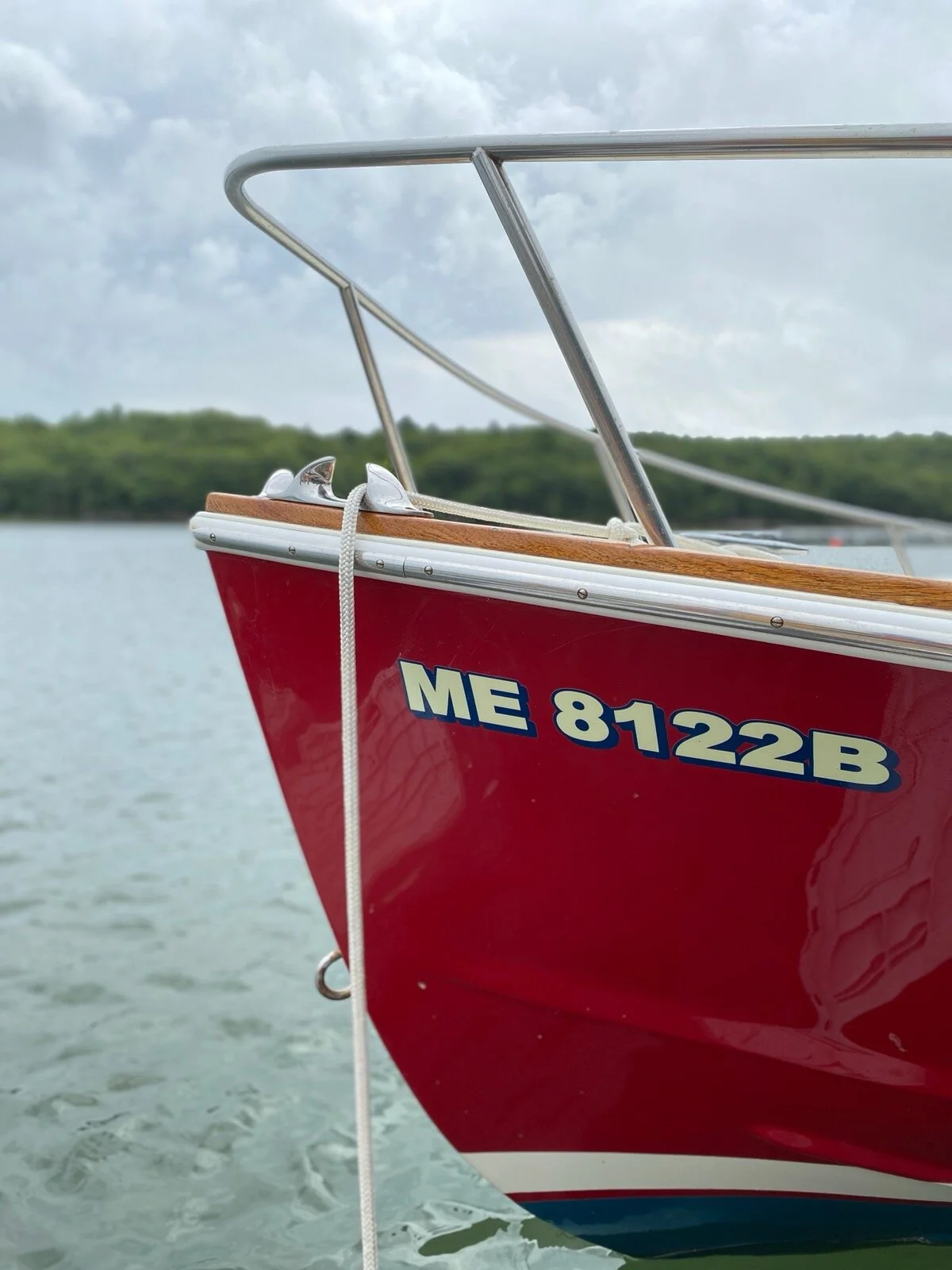 Close-up of the front part of a red boat with boat registration number ME 8122B, floating on a body of water with a distant tree-lined shoreline and cloudy sky in the background.