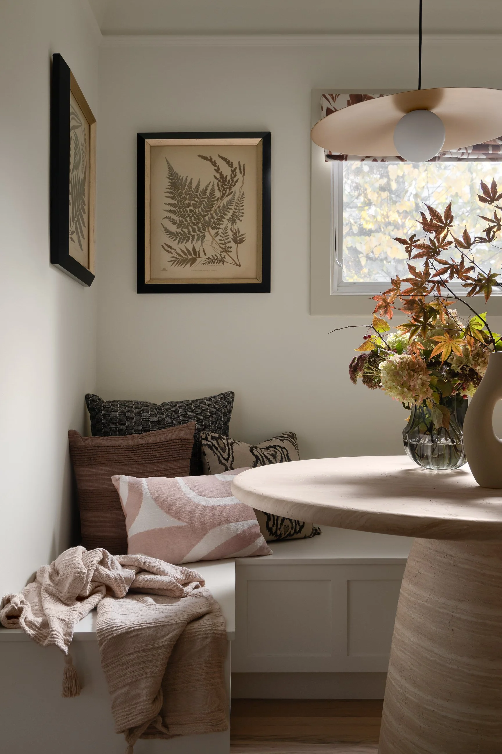 A cozy corner with a window, artwork, and a bouquet of flowers on a light-colored table, with cushions on a window seat and soft blankets.