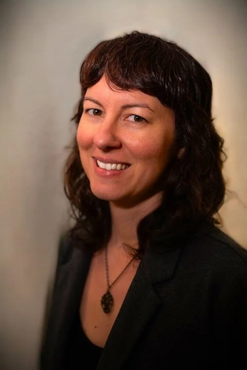 A woman with shoulder-length dark hair, wearing a black blazer and a necklace, smiling at the camera against a plain background.