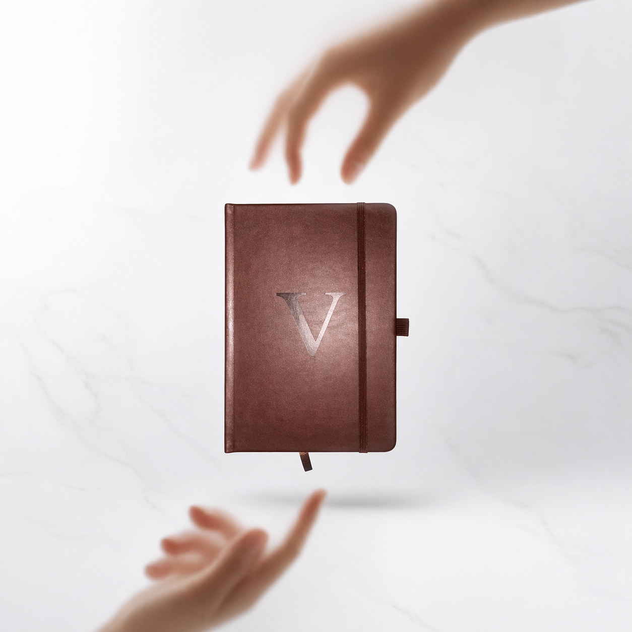 A brown leather-bound journal with a large letter V embossed on the front, floating above a white marble surface with two hands reaching toward it from above and below.
