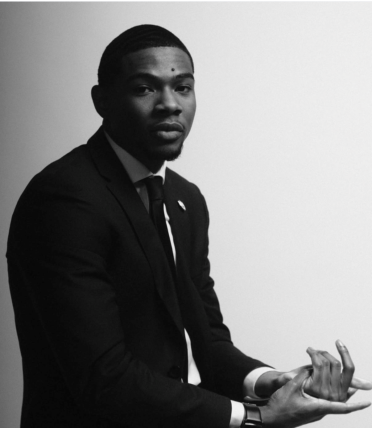 Black and white portrait of a young man in a suit, sitting against a plain background, with a serious expression, looking at the camera, with short hair and a small beard, holding his hands in a gesture.