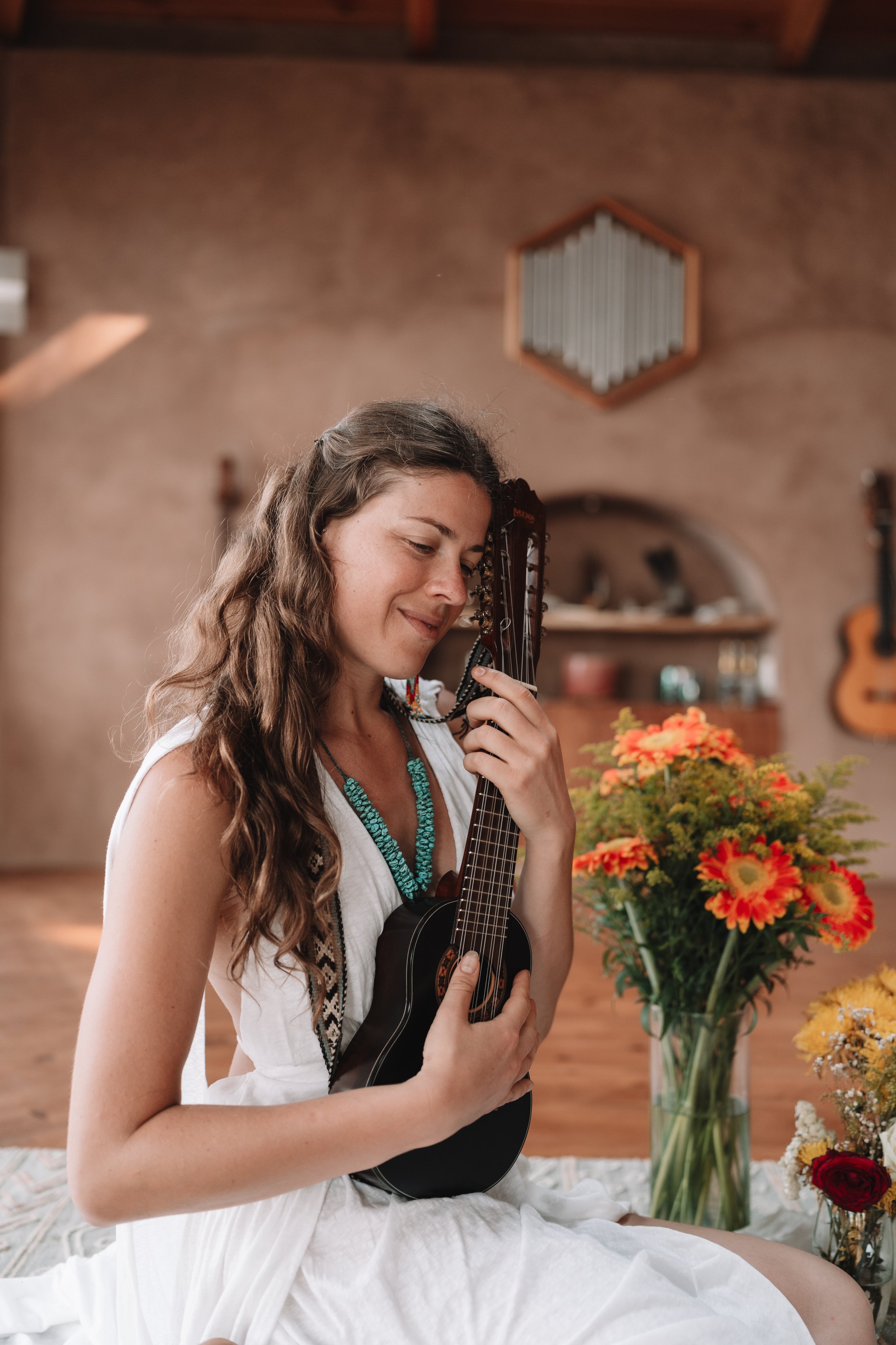 Young woman with long brown hair sitting on floor, playing a black charango, surrounded by colorful flowers in vases, in a cozy room with wooden ceiling and beige walls.