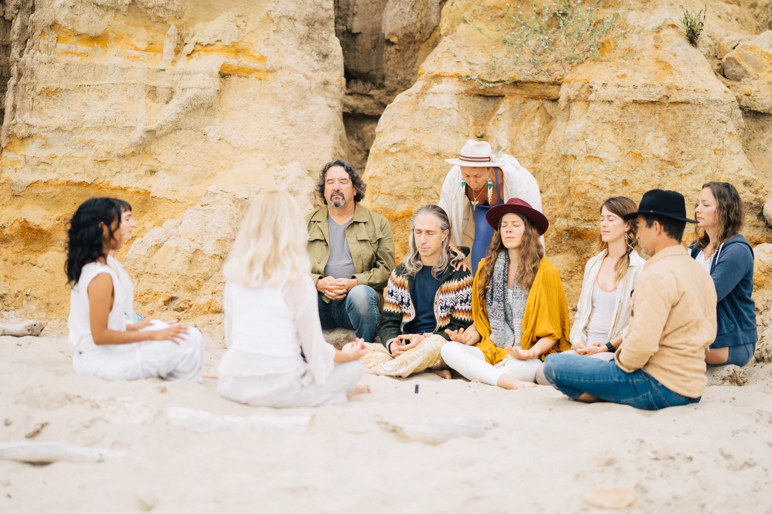 A group of people sitting on the ground, participating in a meditation or spiritual gathering outdoors near rocky cliffs with a woman in white leading.