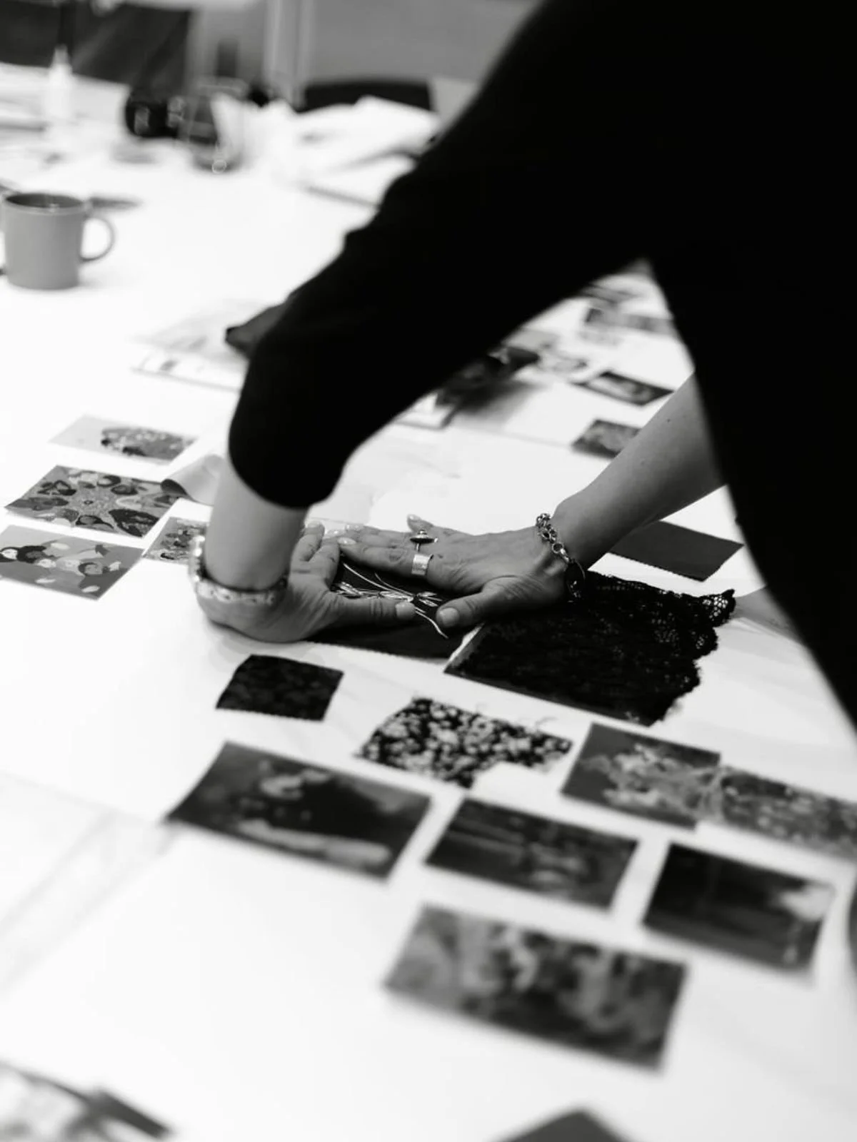 A person is laying out and organizing printed photographs on a table. They are wearing rings and bracelets and are dressed in dark clothing. The table is scattered with various photos, some being held down with their hands, and a coffee mug is visible in the background.