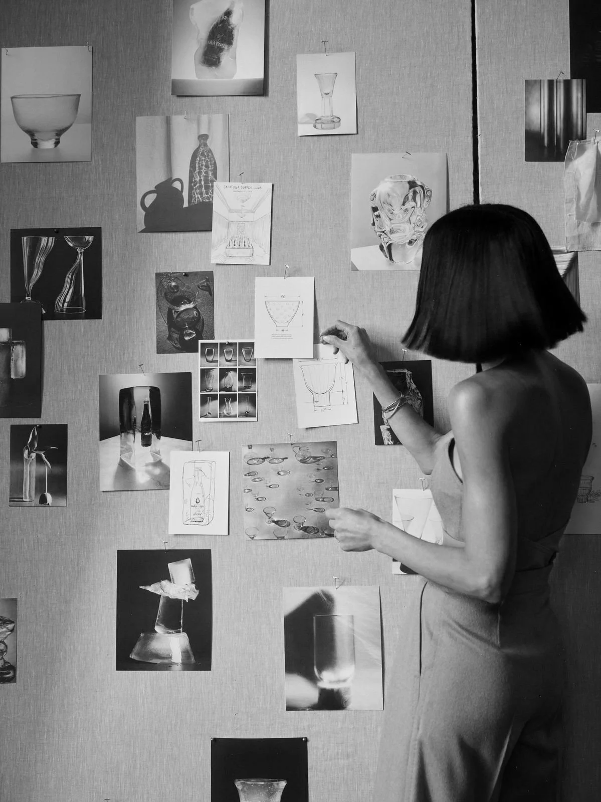 A woman with short dark hair wearing a sleeveless dress is standing in front of a wall covered with various photographs, sketches, and drawings of glassware and bottles, possibly working on or analyzing a design project.