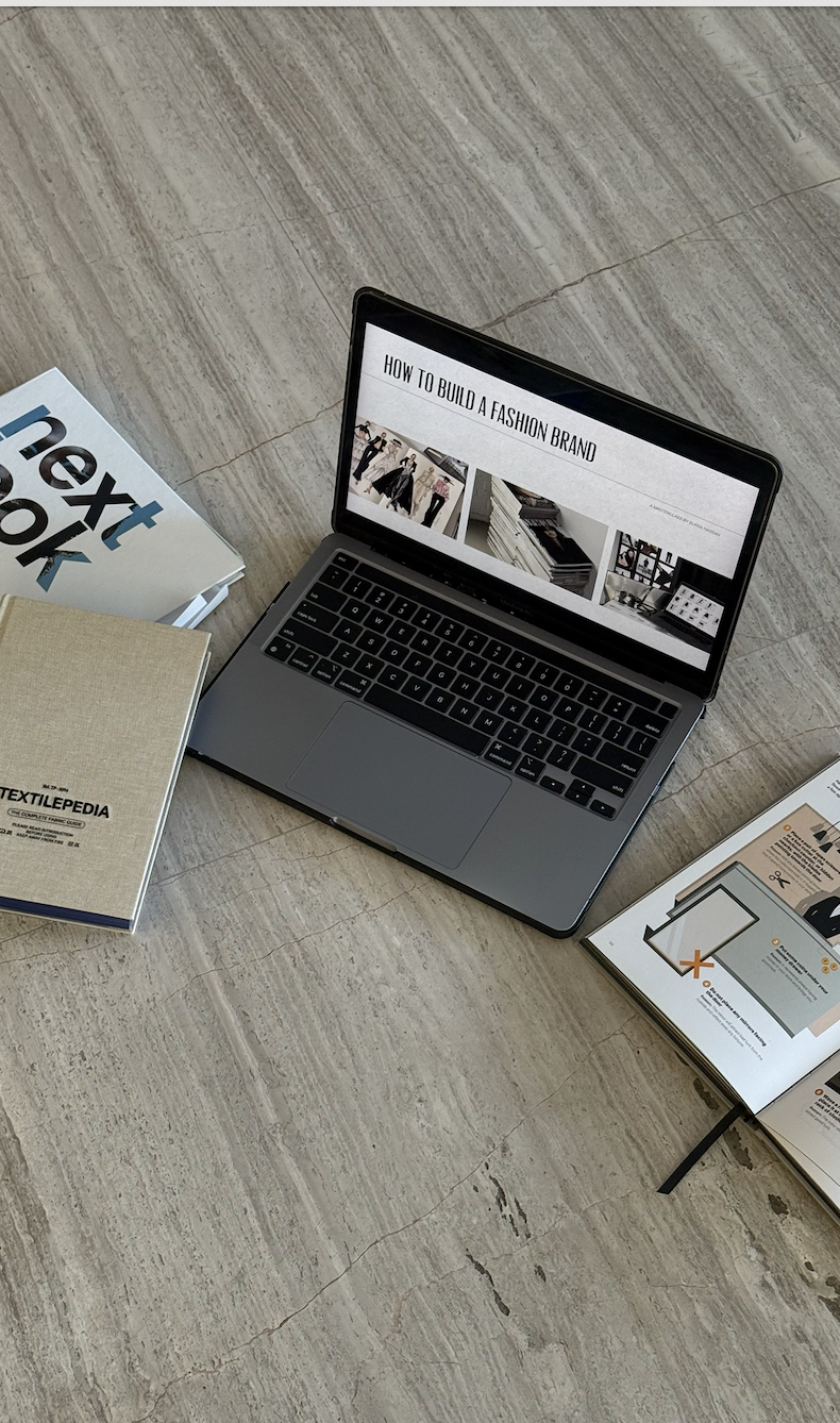 Laptop displaying a presentation titled 'How to Build a Fashion Brand' surrounded by books on fashion and textiles on a marble floor.