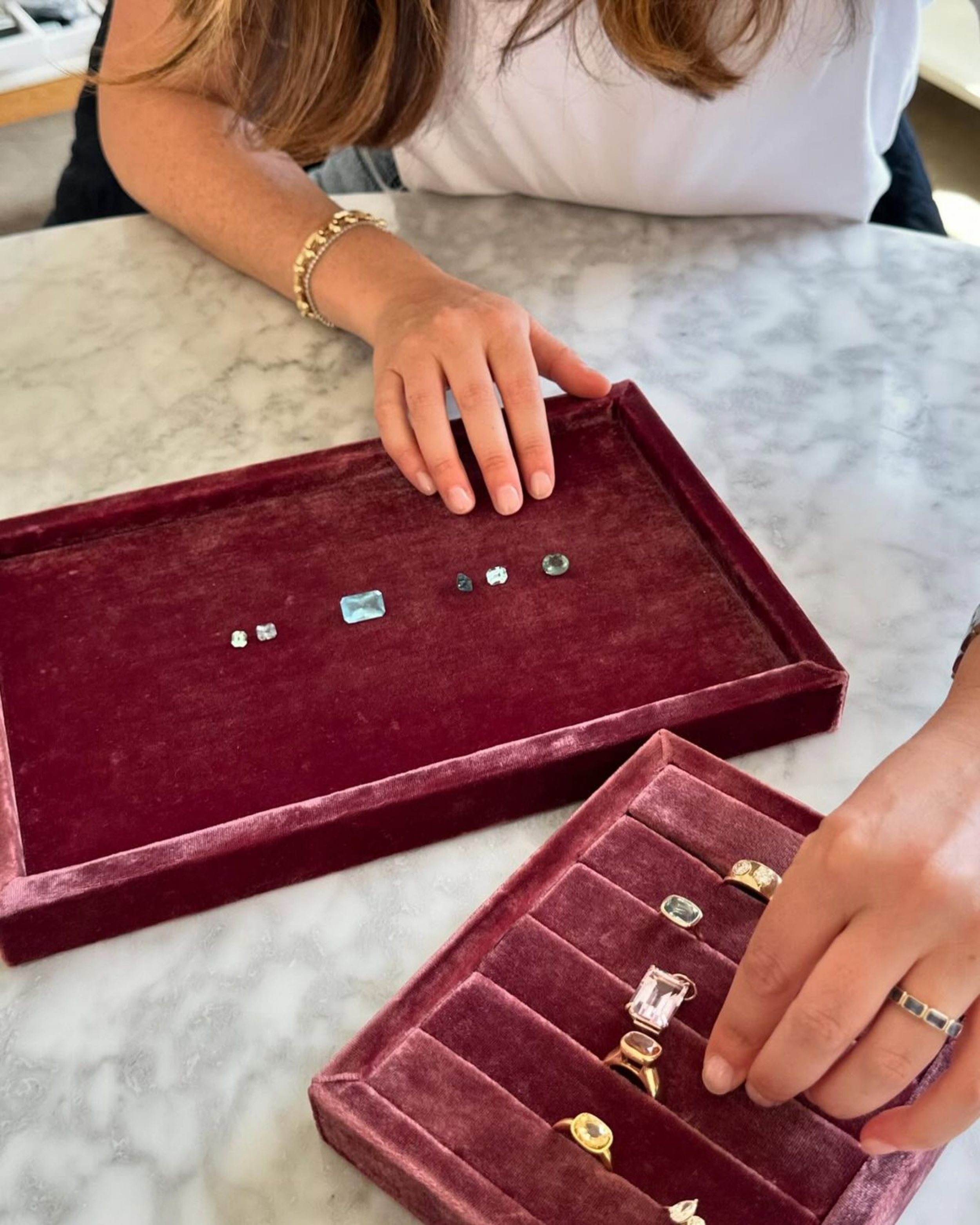 A woman with long reddish-brown hair, wearing a white top and gold jewelry, is placing rings into a purple velvet jewelry box on a white marble surface. The jewelry box contains various rings, some with large gemstones, and others with smaller stones or plain bands.
