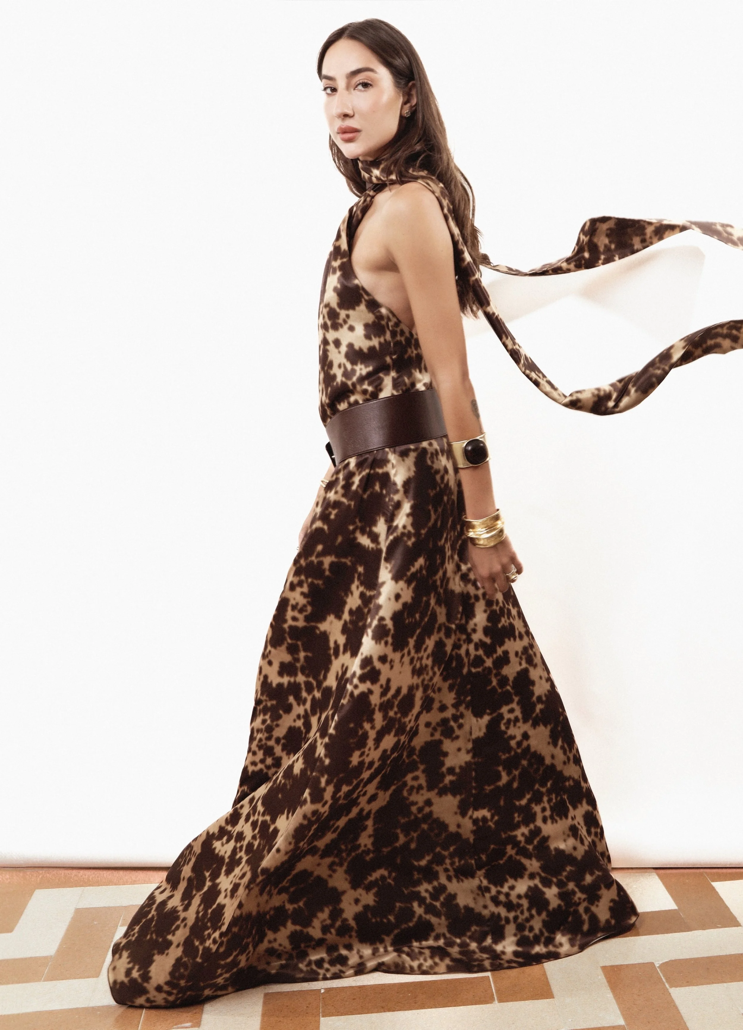 A woman in a leopard print dress with a large matching scarf, standing on a tiled floor against a plain white background.