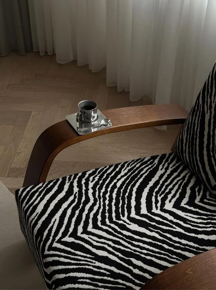 A close-up of an armchair with zebra-striped upholstery and a wooden side table holding a cup of coffee on a tray with a spoon, in front of sheer white curtains.