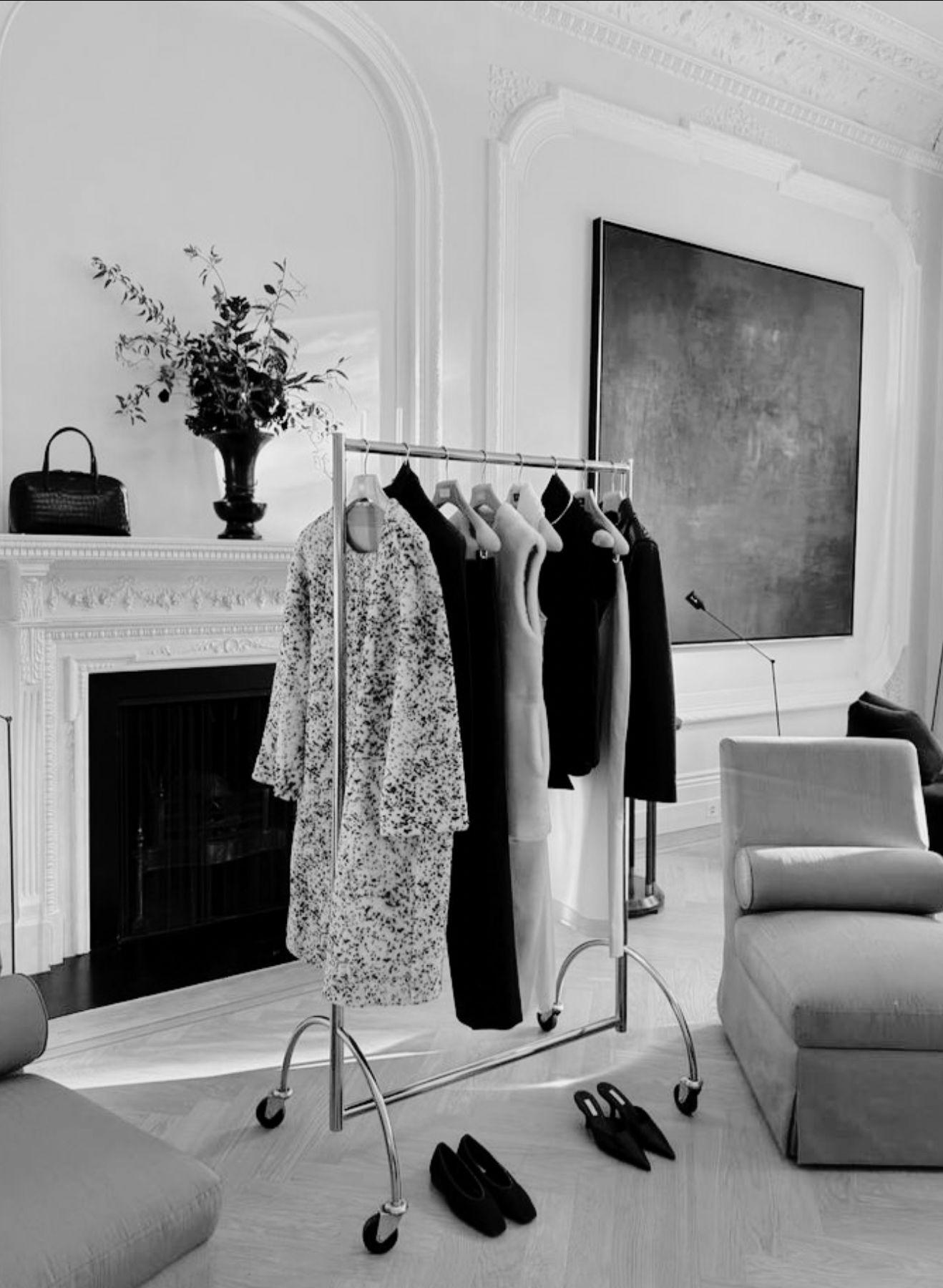 A room with a clothing rack holding dresses and tops, a pair of black shoes on the wooden floor, a white armchair, a fireplace with decorative molding, a large abstract painting, a floor lamp, a table with a flower arrangement and a handbag, and a pair of black shoes next to the outfit rack.