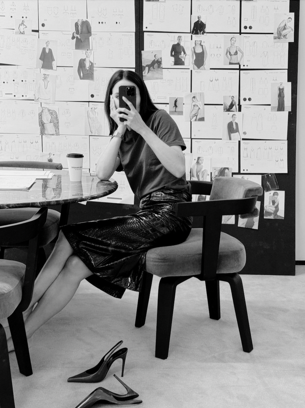 A woman taking a mirror selfie in a room with fashion sketches and photos on the wall, wearing a dark t-shirt, shiny skirt, and high heels placed on the floor.