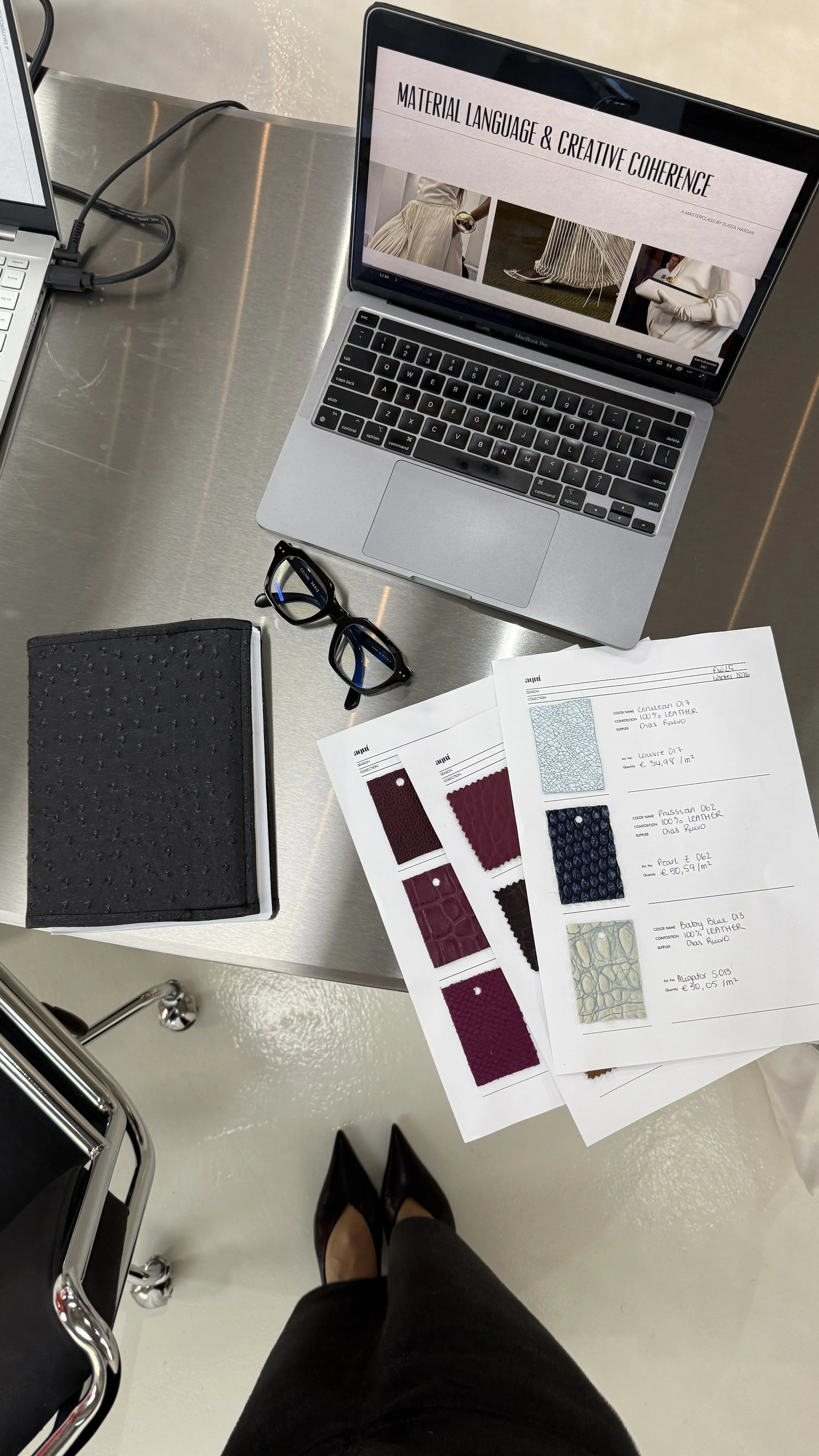 Overhead view of a person's outfit and workspace, including black high heels, black pants, a stainless steel desk, a laptop displaying a presentation titled 'Material Language & Creative Coherence,' a pair of glasses, a black textured notebook, and fabric samples and color charts on printed sheets.
