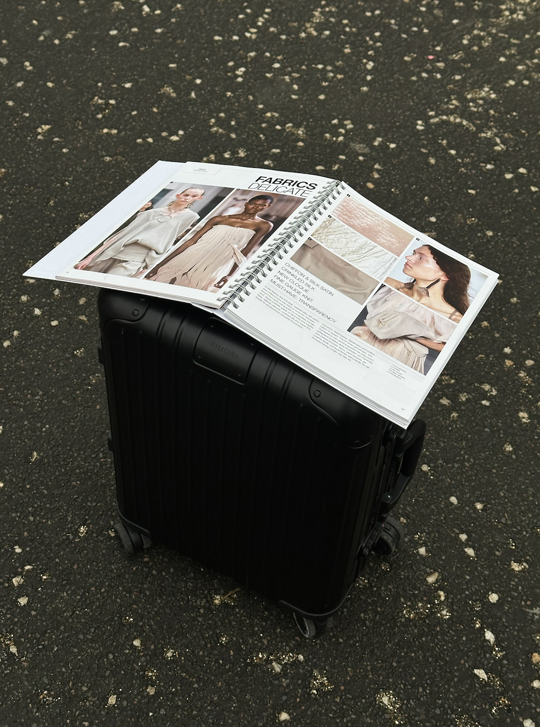 An open fashion catalog resting on a black suitcase on a dark, textured outdoor surface, displaying images and fabric swatches of delicate fabric materials.