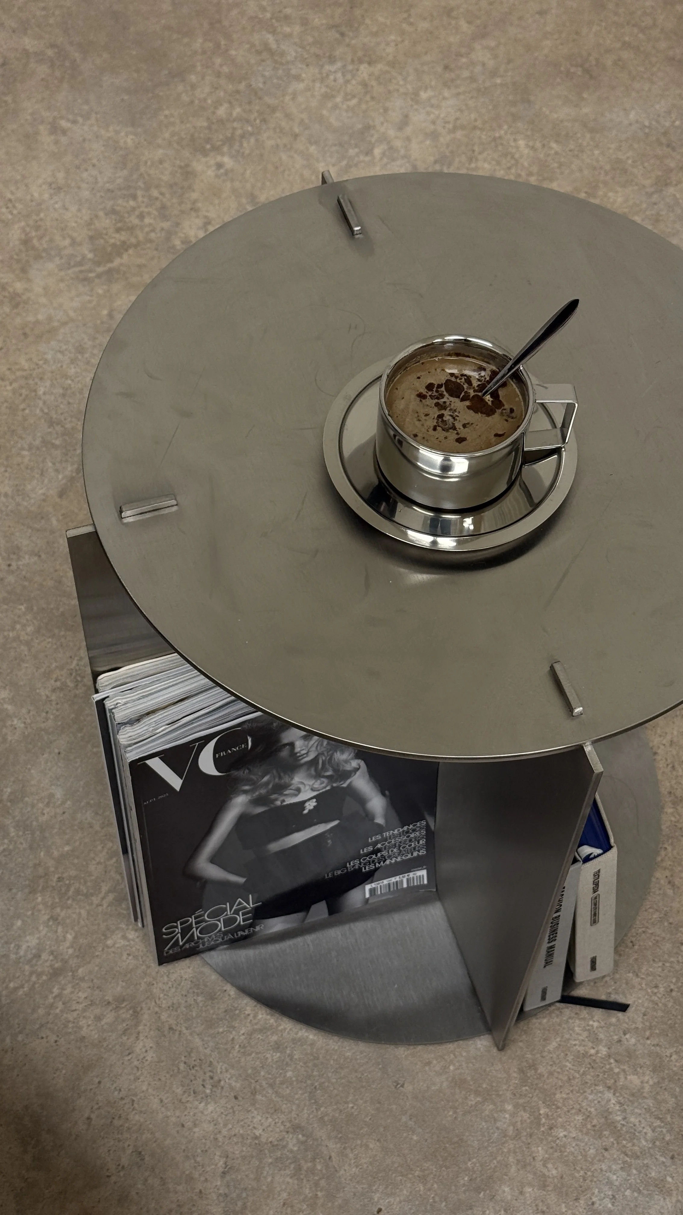 A round, metallic table with a cup of hot chocolate topped with chocolate shavings and a spoon, placed on a small tray. The table also has a magazine or newspaper underneath, featuring a black and white photo of a woman.