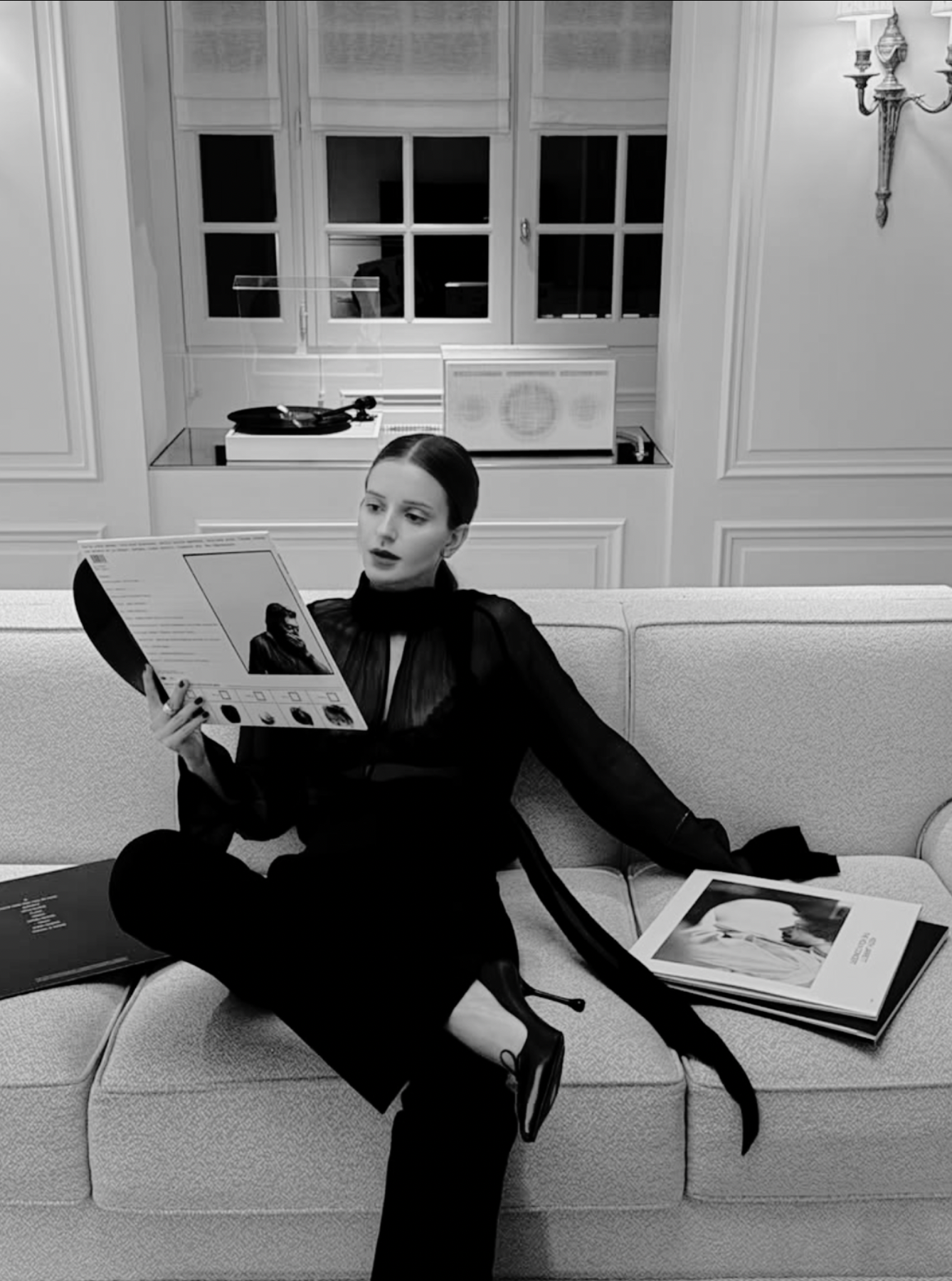 Black and white photo of a woman with dark hair sitting on a sofa, reading a magazine. She is dressed in a dark, sheer blouse with a high collar and long sleeves, combined with dark pants and high-heeled boots. There are multiple magazines on the sofa around her. The room has a window with blinds in the background and an ornate wall sconce on the right side, with decorative paneling on the wall.