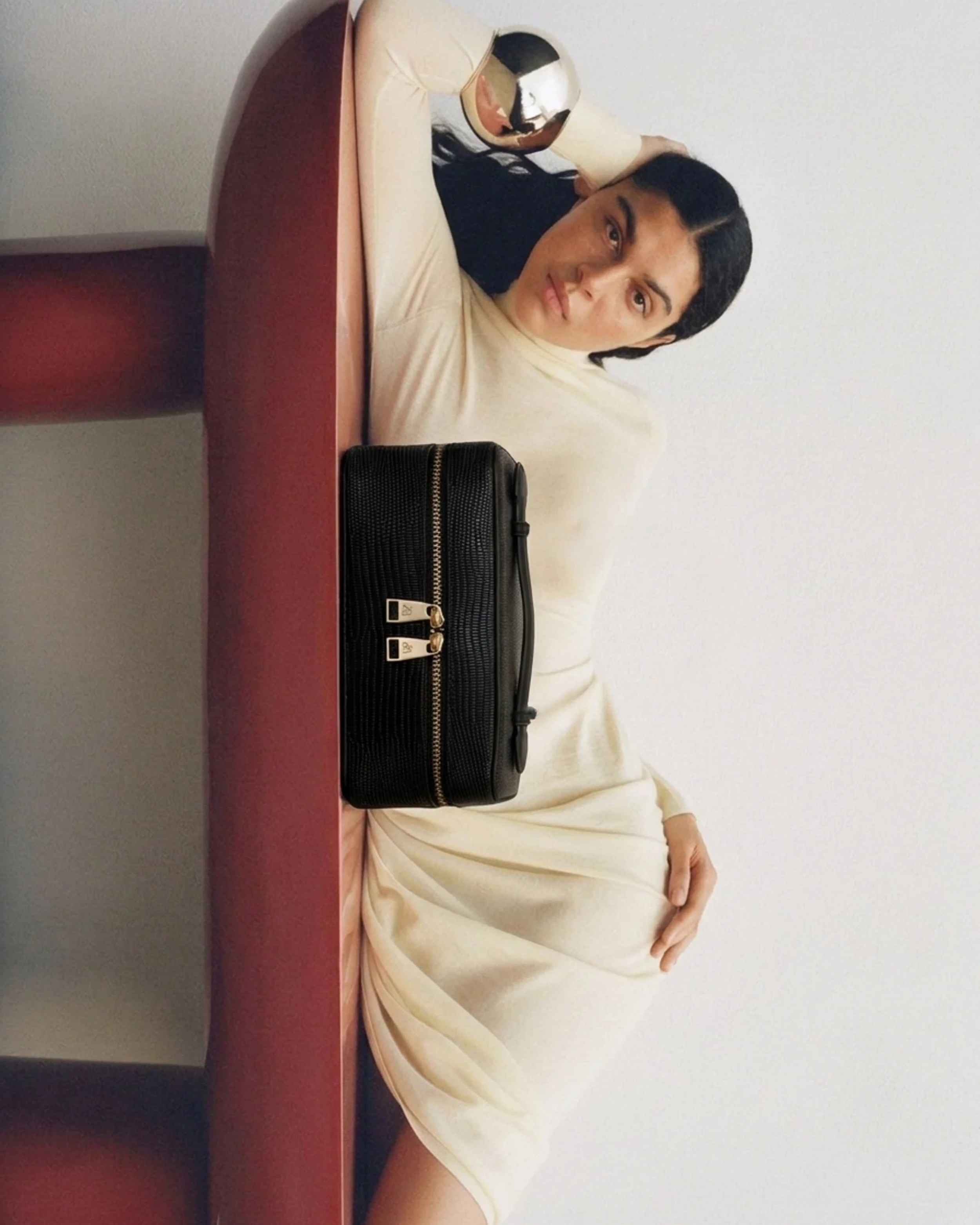 A woman with dark wavy hair and light skin is lying on a bed or couch, resting her head on her arm. She is wearing a beige-colored dress and is looking directly at the camera with a neutral expression. There is a black purse with gold accents placed on the bed in front of her and a metallic gold and black object, possibly a speaker or a decorative item, beside her. The background is plain and white, and the image is taken indoors.