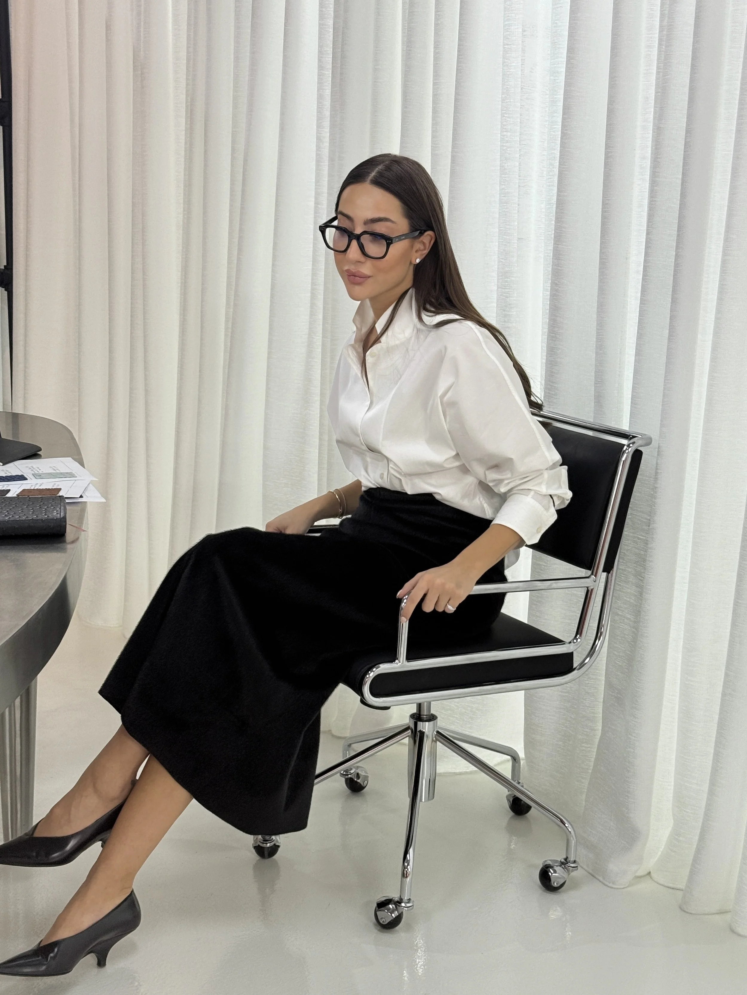 A woman with long dark hair, wearing glasses, a white blouse, and a black skirt, sitting on a black office chair with chrome arms in front of a white curtain. She is dressed professionally and appears to be in an office setting.