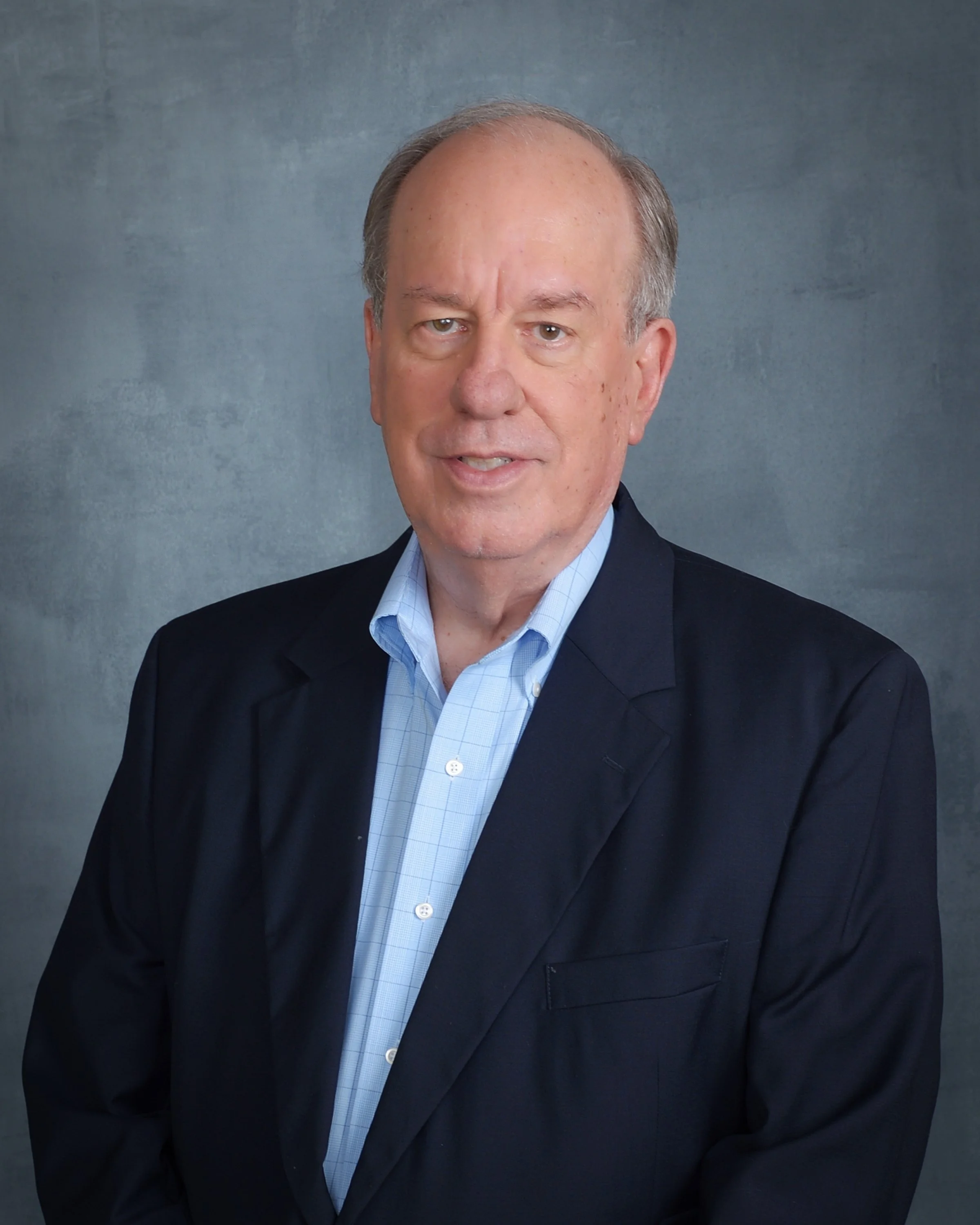 A middle-aged man , wearing a blue blazer over a light blue button-up shirt, standing against a plain background.