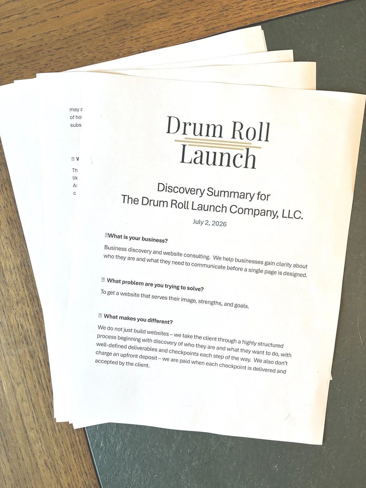 A printed document titled "Drum Roll Launch" with a subtitle "Discovery Summary for The Drum Roll Launch Company, LLC." , lying on top of multiple sheets of paper on a wooden table. The document contains headings and text about business discovery and website consulting.