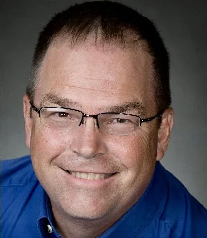A man with glasses wearing a blue shirt smiling at the camera.