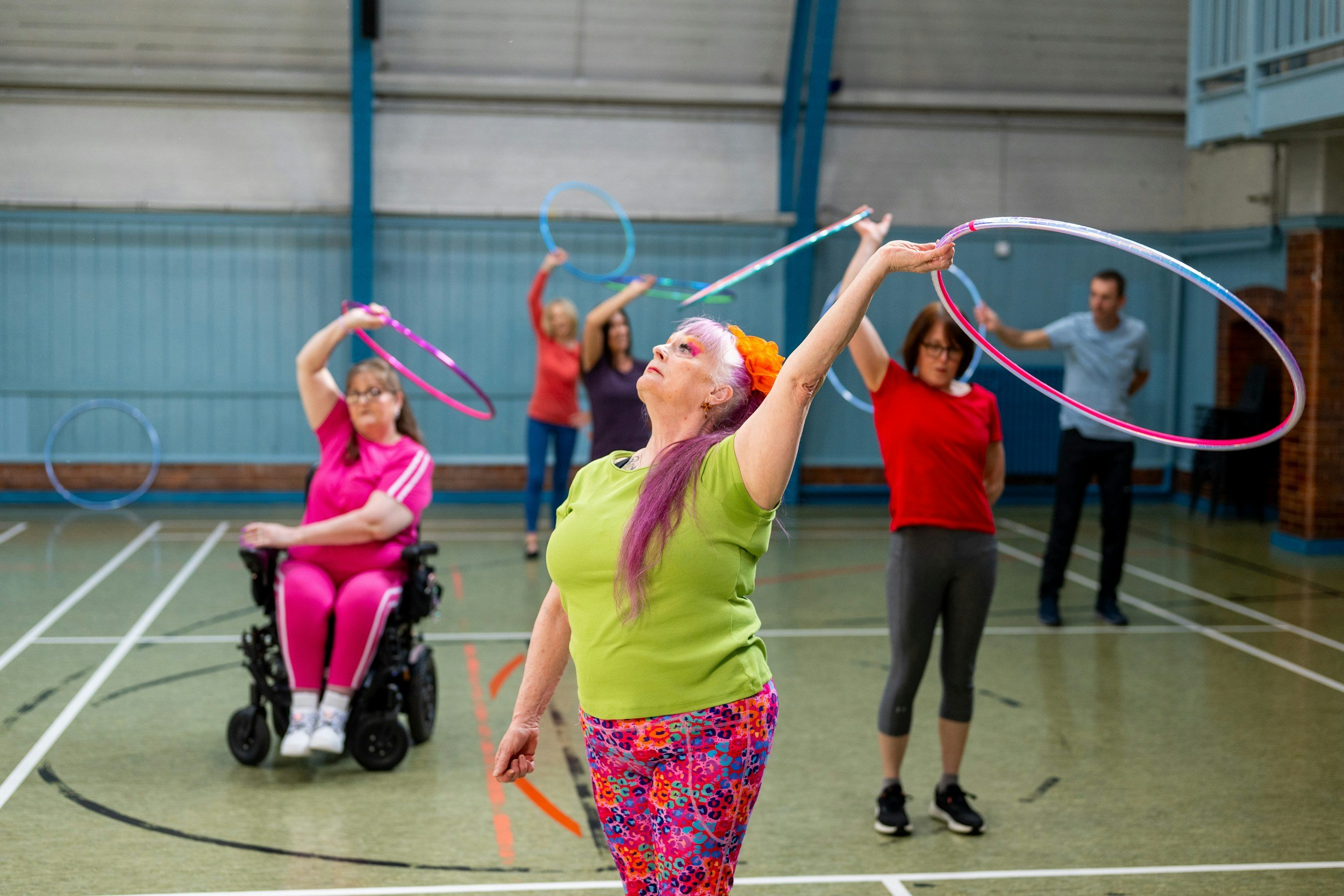 A diverse group of people, including a woman in a wheelchair, participating in a hula hoop class indoors. The woman in the foreground has purple hair and colorful leggings, and is skillfully spinning a hula hoop around her waist. Others in the background are also engaged in hula hooping and appear to be enjoying the activity.