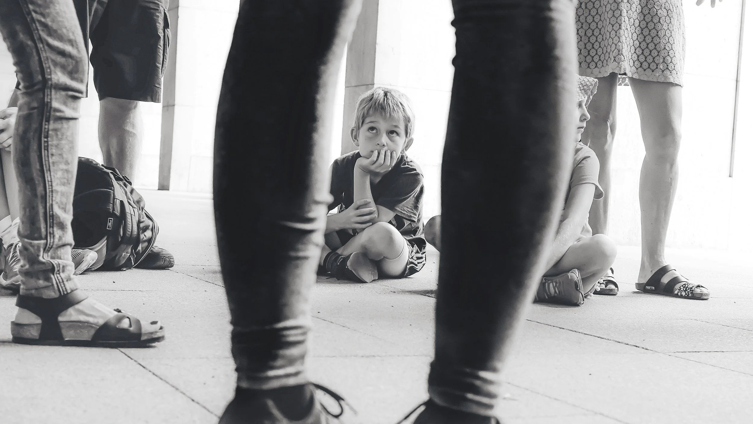A young boy sitting on the floor, looking up pensively through the legs of standing people. The scene appears to be indoors, with the boy surrounded by adults.