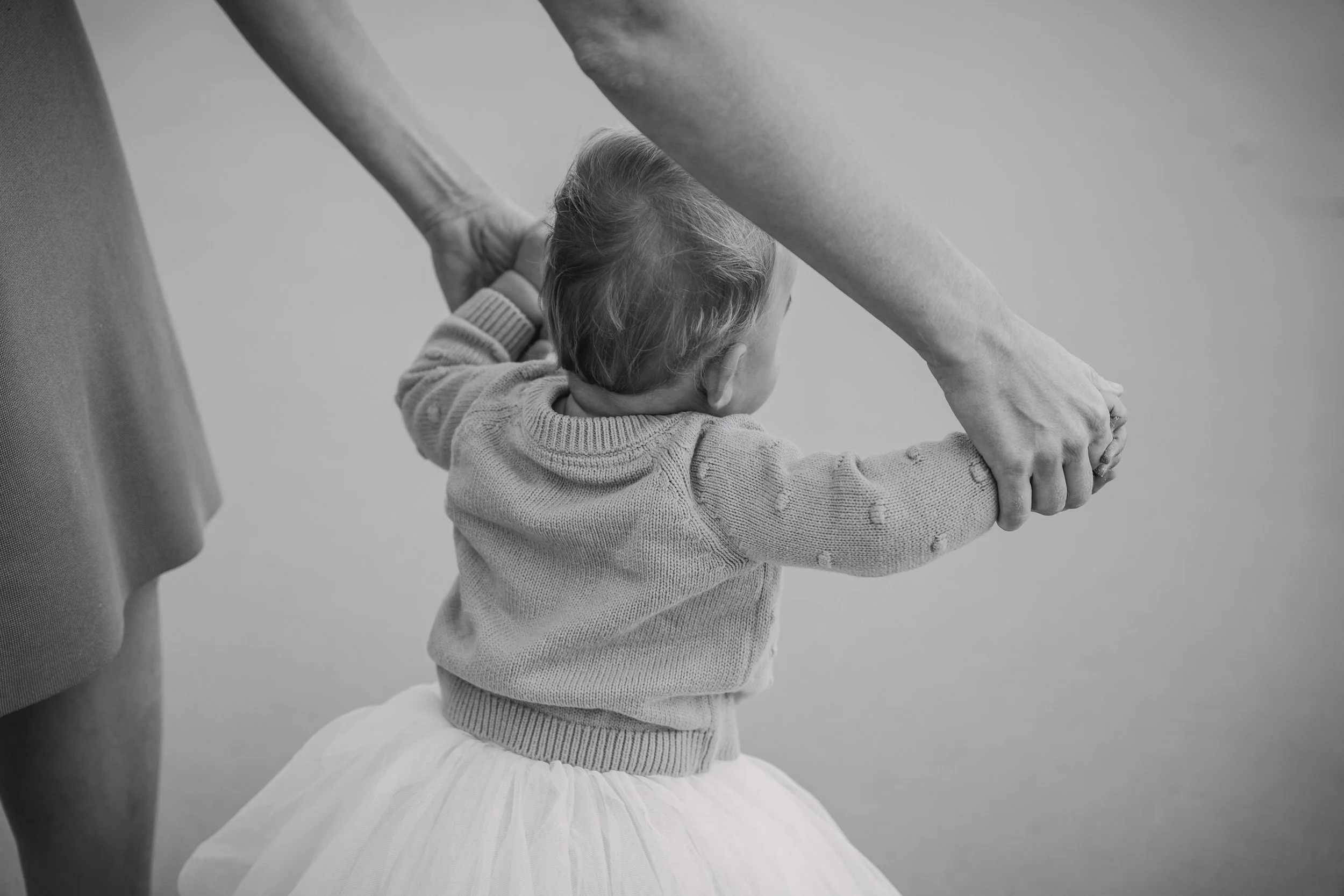 A young girl practicing ballet with an adult in a dance studio, the girl is dressed in a sweater and tutu, and is holding her head with her hands while standing on her knees.