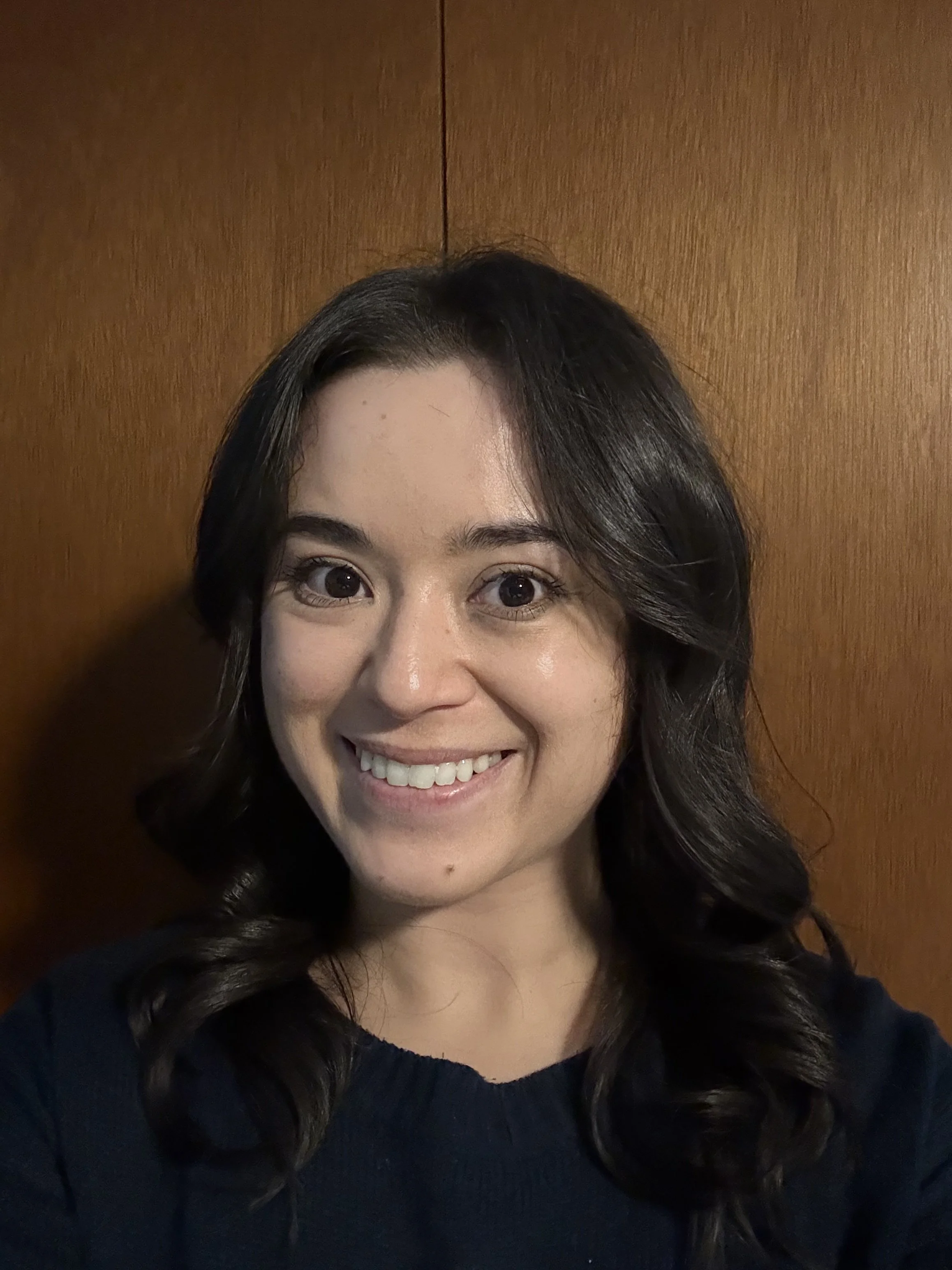 Close-up of a smiling woman with dark, wavy hair against a wooden background.