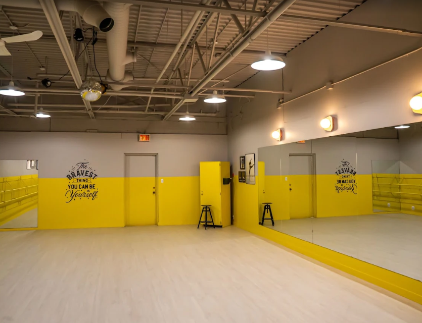 A brightly lit dance studio with yellow and white walls, a large mirror, and wooden flooring. Motivational quote on the wall reads, 'The bravest thing you can be is yourself.'