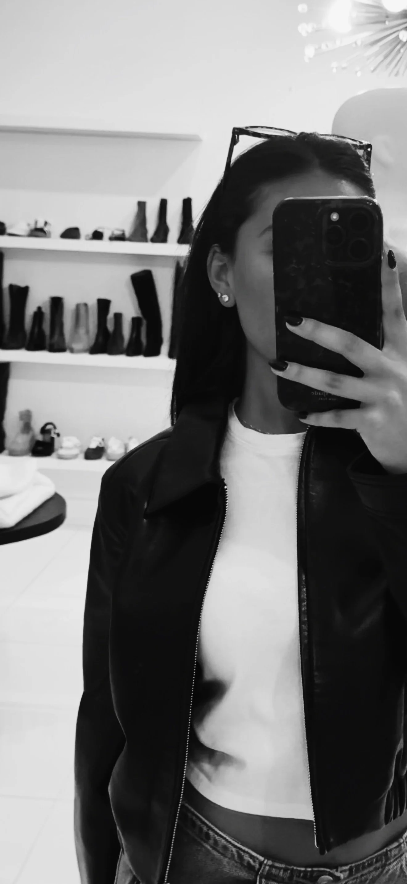 Black and white photo of a woman taking a mirror selfie with her phone, showing her face mostly covered by the phone. She is wearing earrings, sunglasses on her head, a leather jacket, and a white shirt. In the background, there are shelves with various bottles and containers.