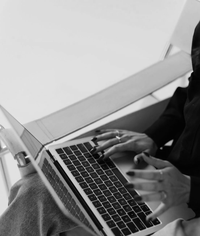 A person typing on a laptop keyboard, with hands resting on the device and wearing rings, in a monochrome setting.