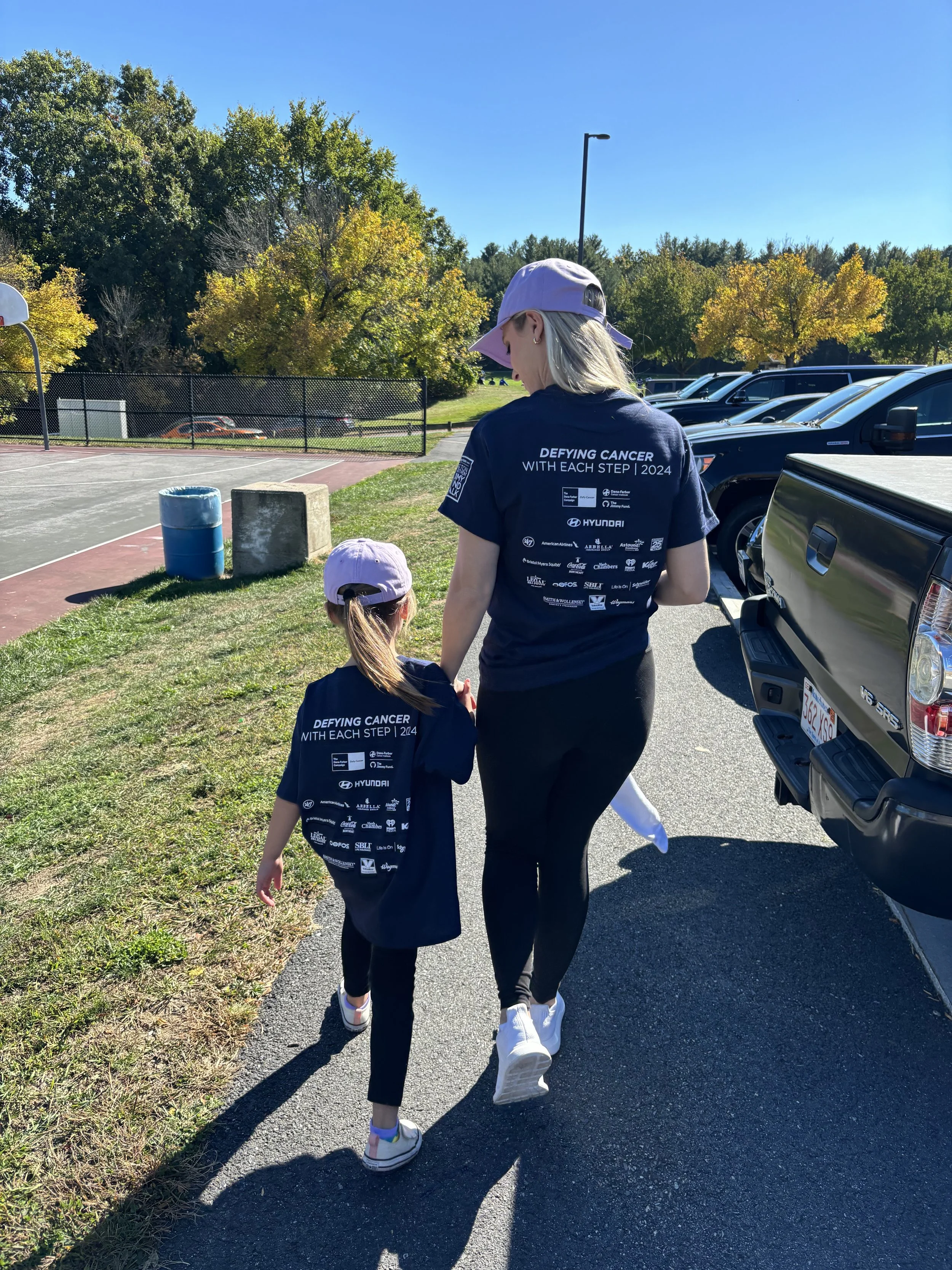 A woman and a young girl walk together outdoors near a parking lot, wearing matching dark shirts with a cancer awareness message on the back, under a clear blue sky.
