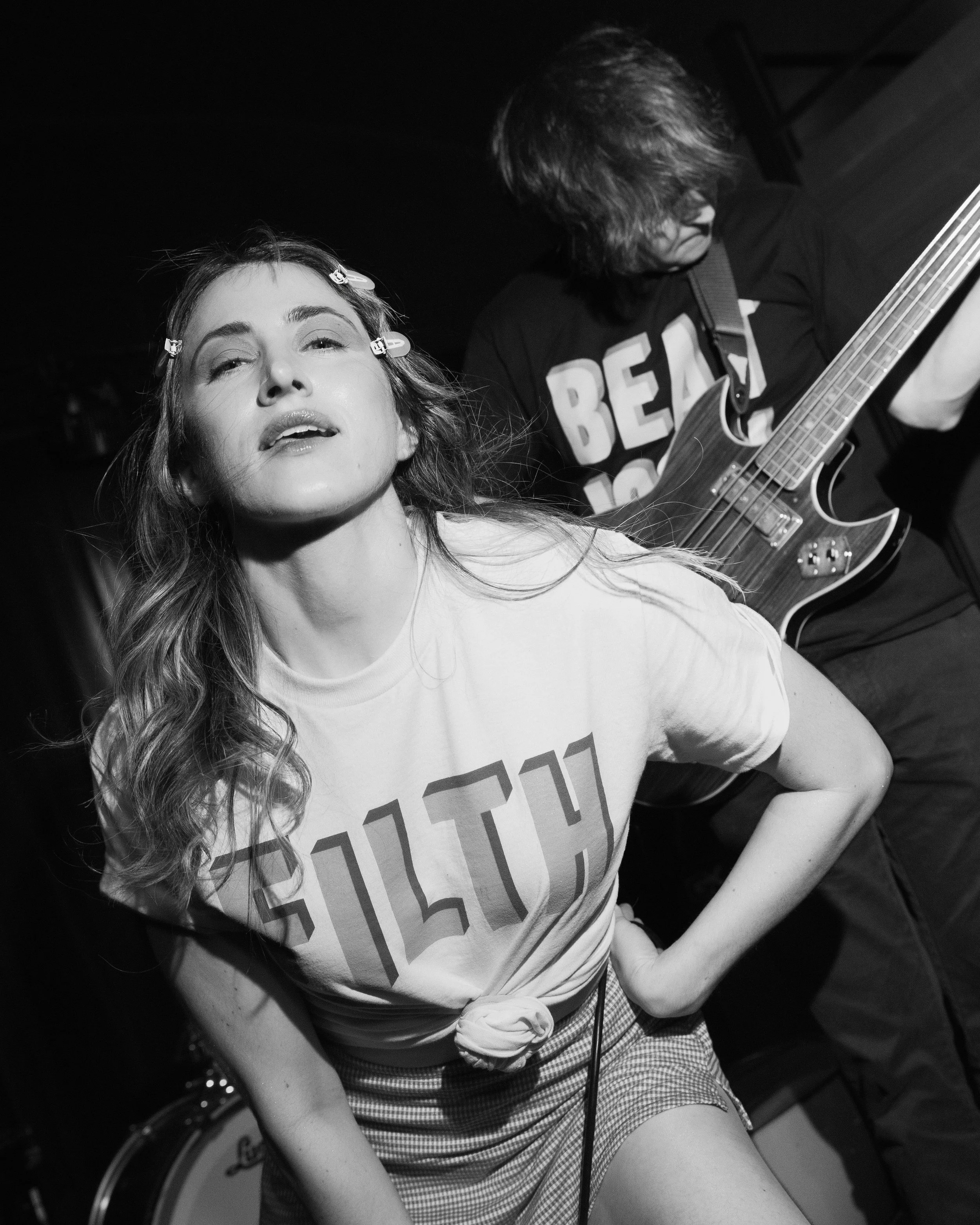 A woman with long hair and hairpins, wearing a T-shirt with large letters, is sitting with her arm resting on a surface, while a person with longer, slightly wavy hair is playing an electric guitar in a dark room.