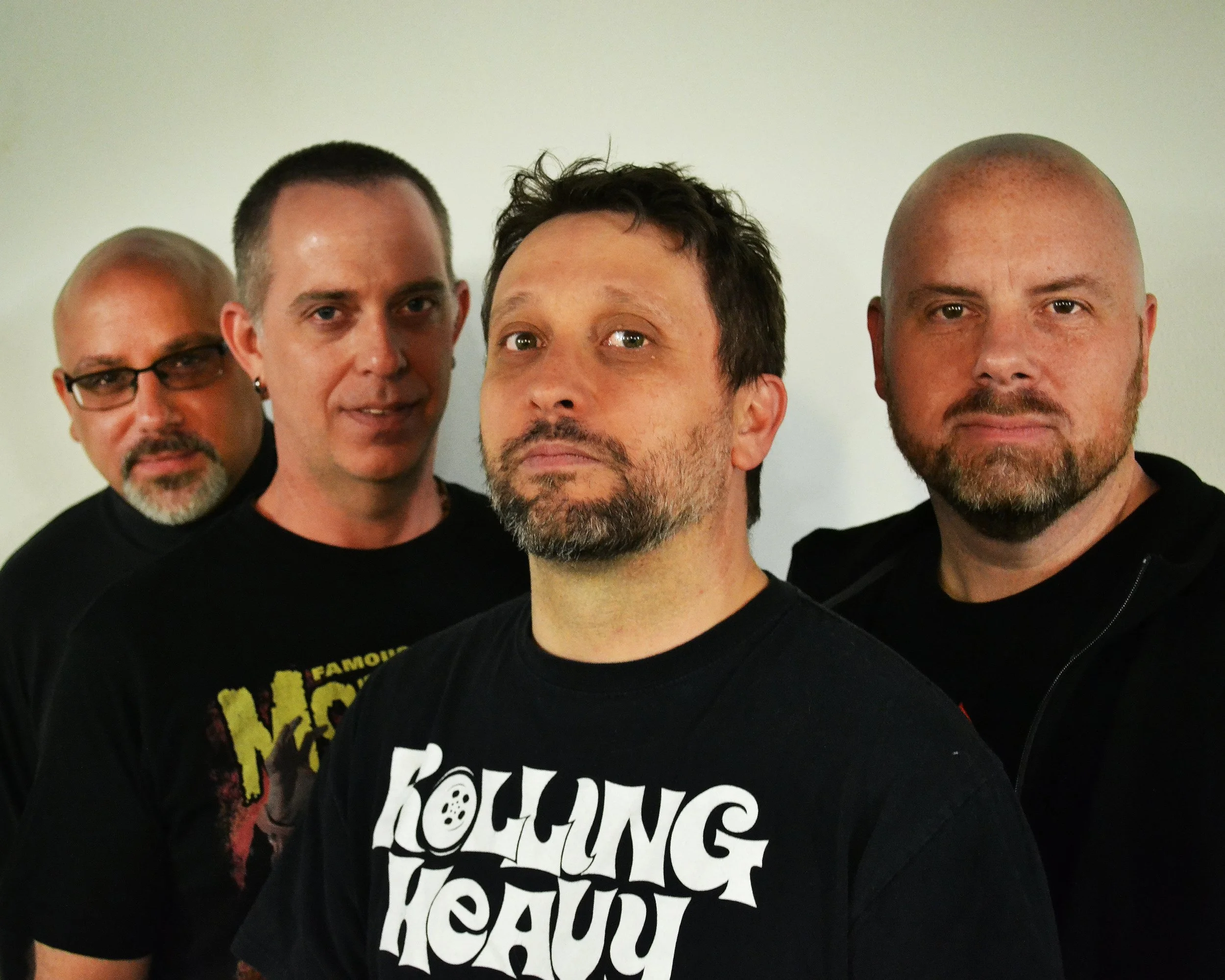 Close-up of four men standing in front of a plain wall, wearing black shirts, with expressions varying from serious to neutral.