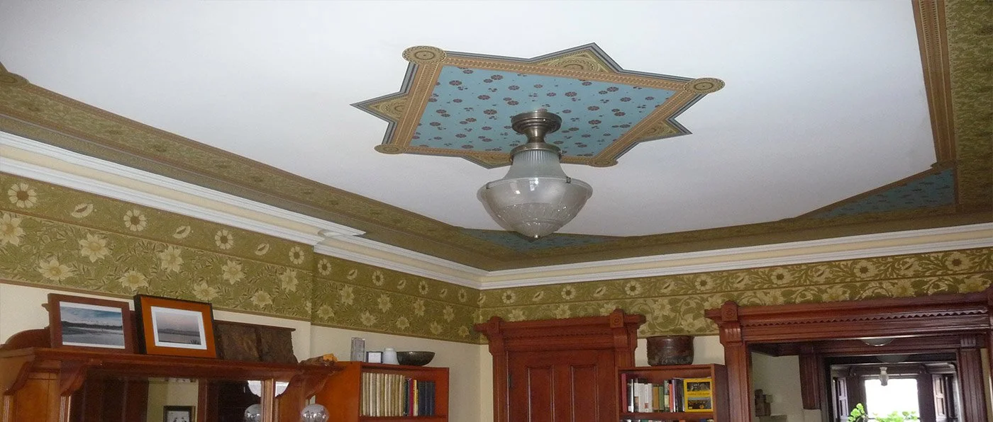 Interior shot of a vintage living room ceiling with ornate crown molding, floral wallpaper border, and a decorative painted ceiling panel. A glass light fixture hangs from the ceiling.