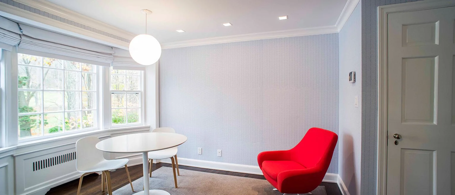 Living room with large windows, white round dining table and chairs, a red armchair, and a white door.