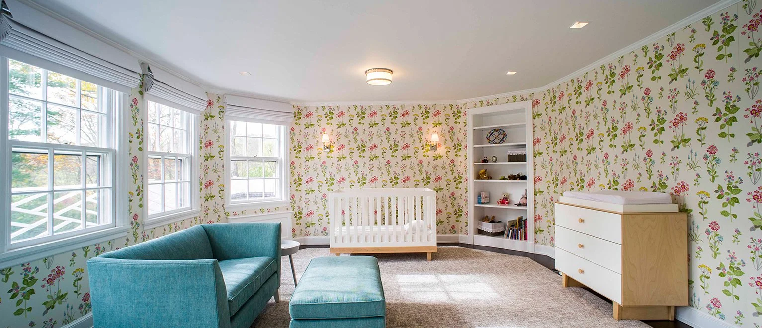 A nursery room with floral wallpaper, a blue sofa with a matching ottoman, a white crib, built-in shelves with toys and books, and a changing table.