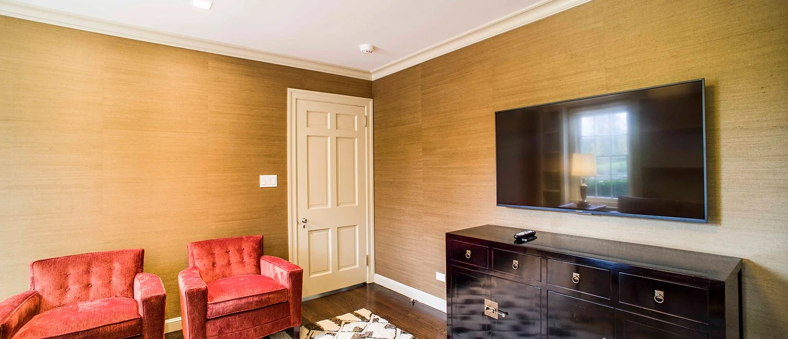 Living room with beige textured walls, a white door, a flat-screen TV mounted on the wall, a dark wood cabinet beneath the TV, a remote control on the cabinet, a window with curtains reflecting in the TV, two red upholstered armchairs, and a rug on t