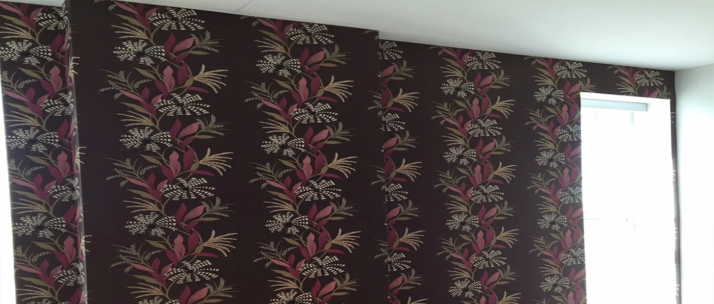 A wall with dark-colored wallpaper featuring a pattern of pink, beige, and white leaves and branches, with a window on the right side letting in natural light.
