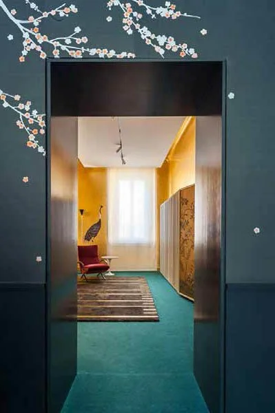 View of a hallway with yellow walls and a window at the end, framed by an opening with dark blue walls decorated with floral artwork.