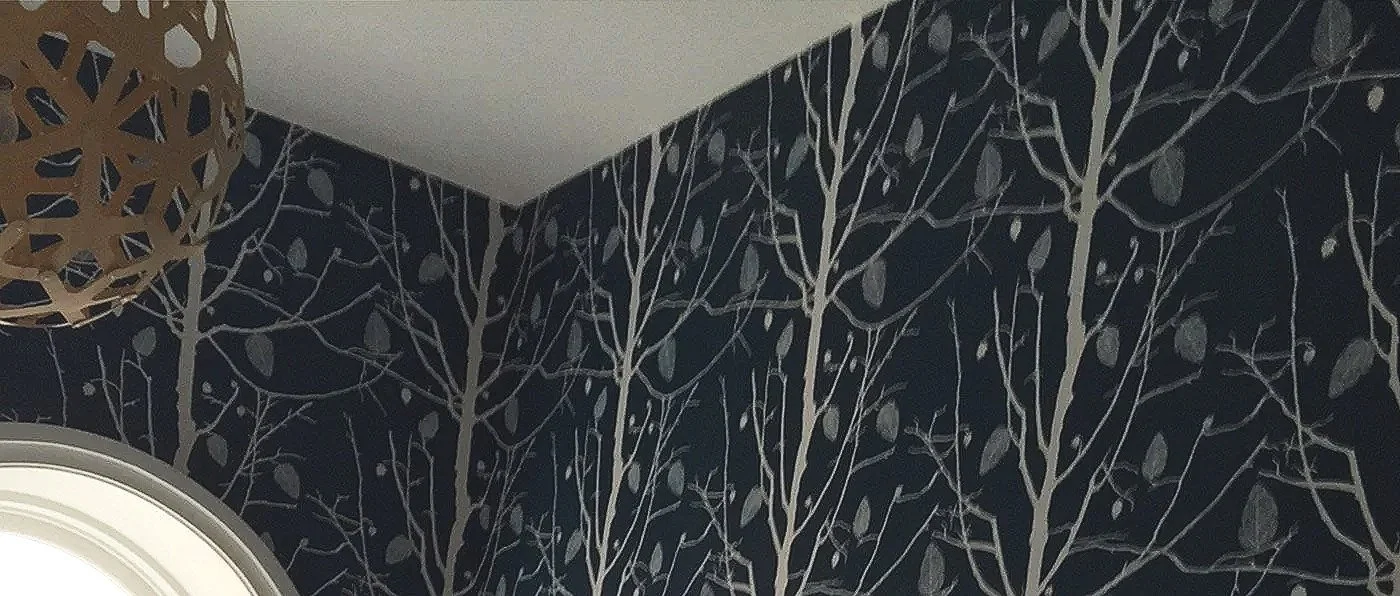 Close-up of a dark navy wallpaper with a pattern of white tree branches and leaves, round ceiling light fixture, and a corner of a window.