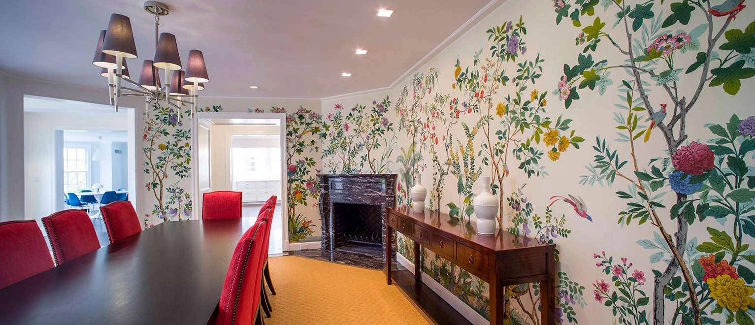 Dining room with a black chandelier, red upholstered chairs around a dark wood table, a yellow rug, and a decorative floral wallpaper wall behind a fireplace and a wooden sideboard with white vases.