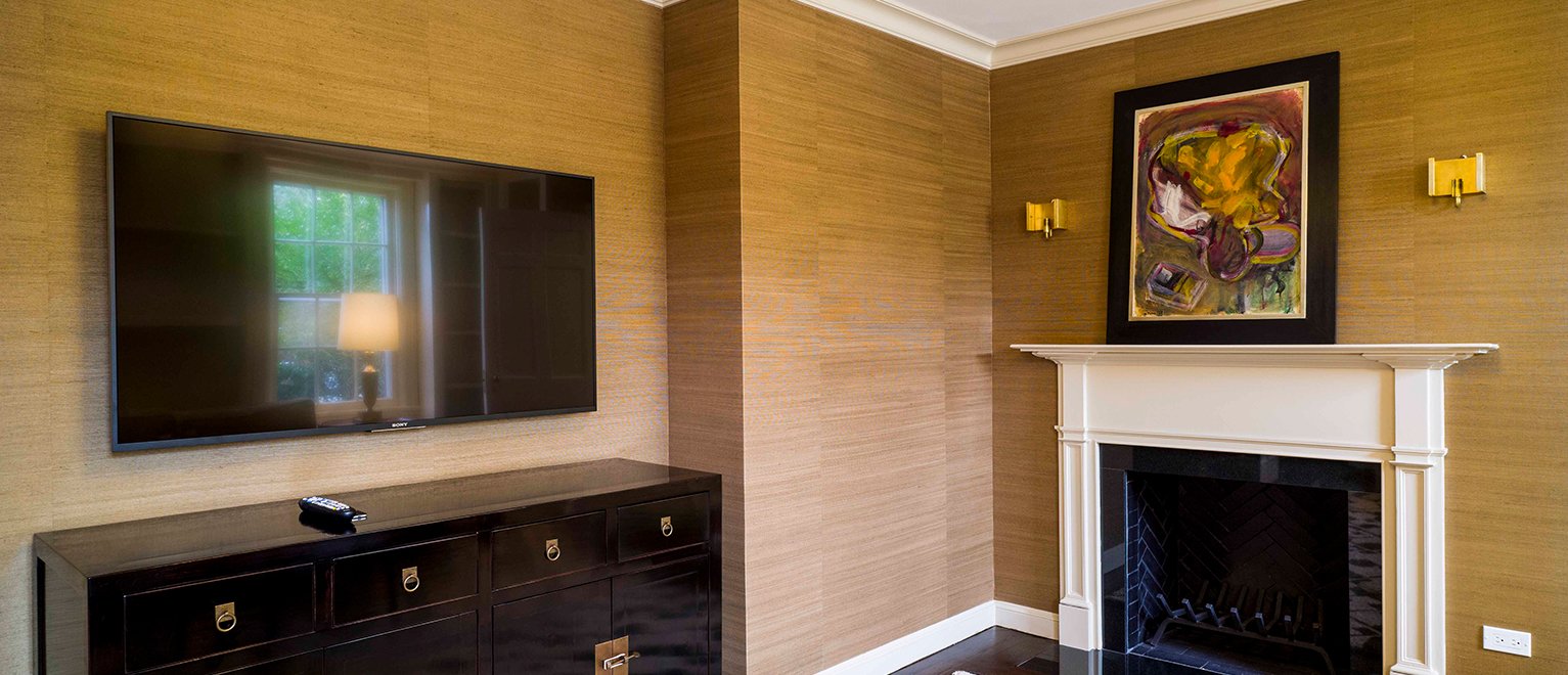 Living room with a wall-mounted TV above a black cabinet, a white fireplace with a colorful abstract painting above, and two gold wall sconces on a beige wall.