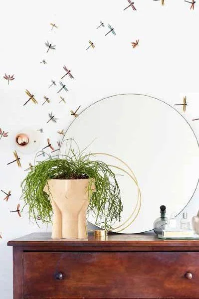 Decorative wall with dragonfly motifs, a large circular mirror, a potted plant, and decorative items on a wooden surface.