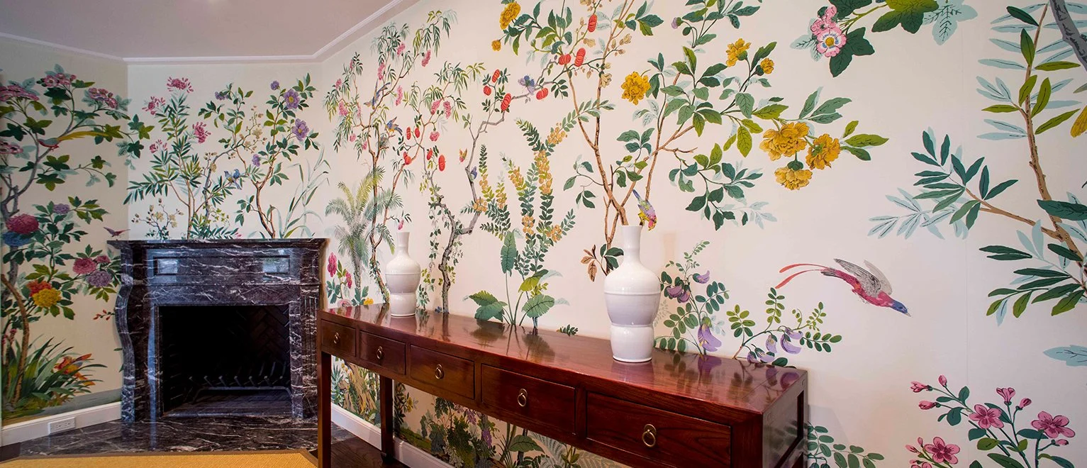 Interior of a room with a colorful botanical wall mural featuring various plants and a bird, a dark marble fireplace, and a wooden sideboard decorated with three white vases.