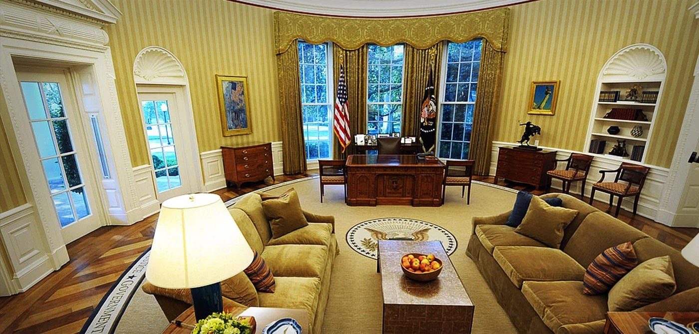 The office features two beige sofas with pillows, a wooden coffee table with a bowl of apples, a large wooden desk with a chair behind it, flanked by American flags, and various artwork and furniture along the walls, with large windows and yellow-striped wallpaper.